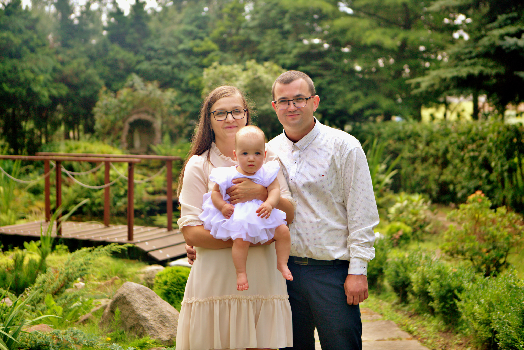 Portret rodziny z małym dzieckiem w białej sukience, na tle parkowej scenerii z mostkiem i zielenią.