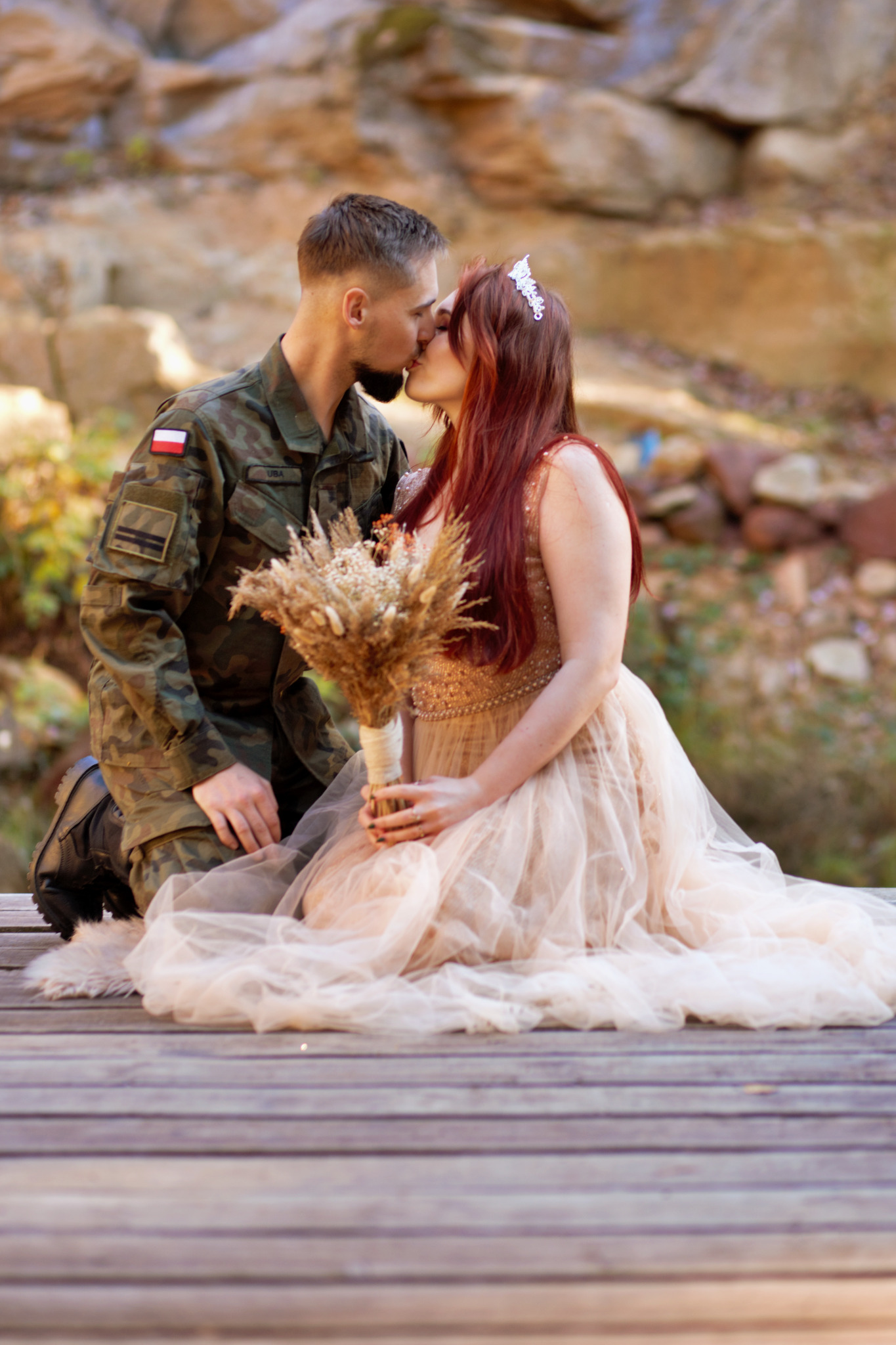 Para całująca się na drewnianym pomoście: mężczyzna w mundurze Wojska Polskiego, kobieta w zwiewnej sukni z bukietem suszonych kwiatów i diademem na rudych włosach, tło z kamieniami.