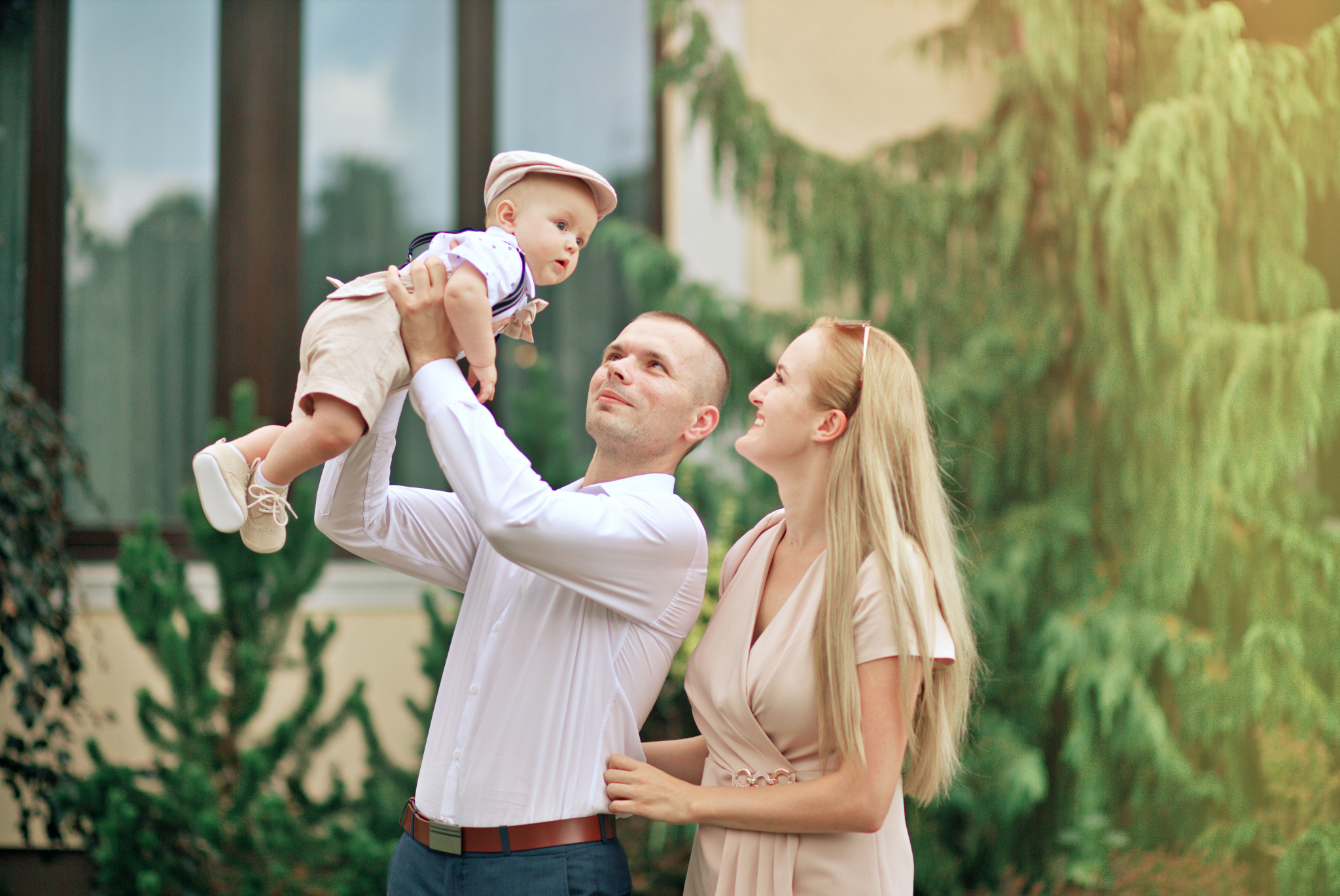 Uśmiechnięta rodzina: ojciec trzyma podniesionego w górę synka w beżowym ubranku i kaszkiecie, matka patrzy z radością, w tle zielone krzewy i budynek.