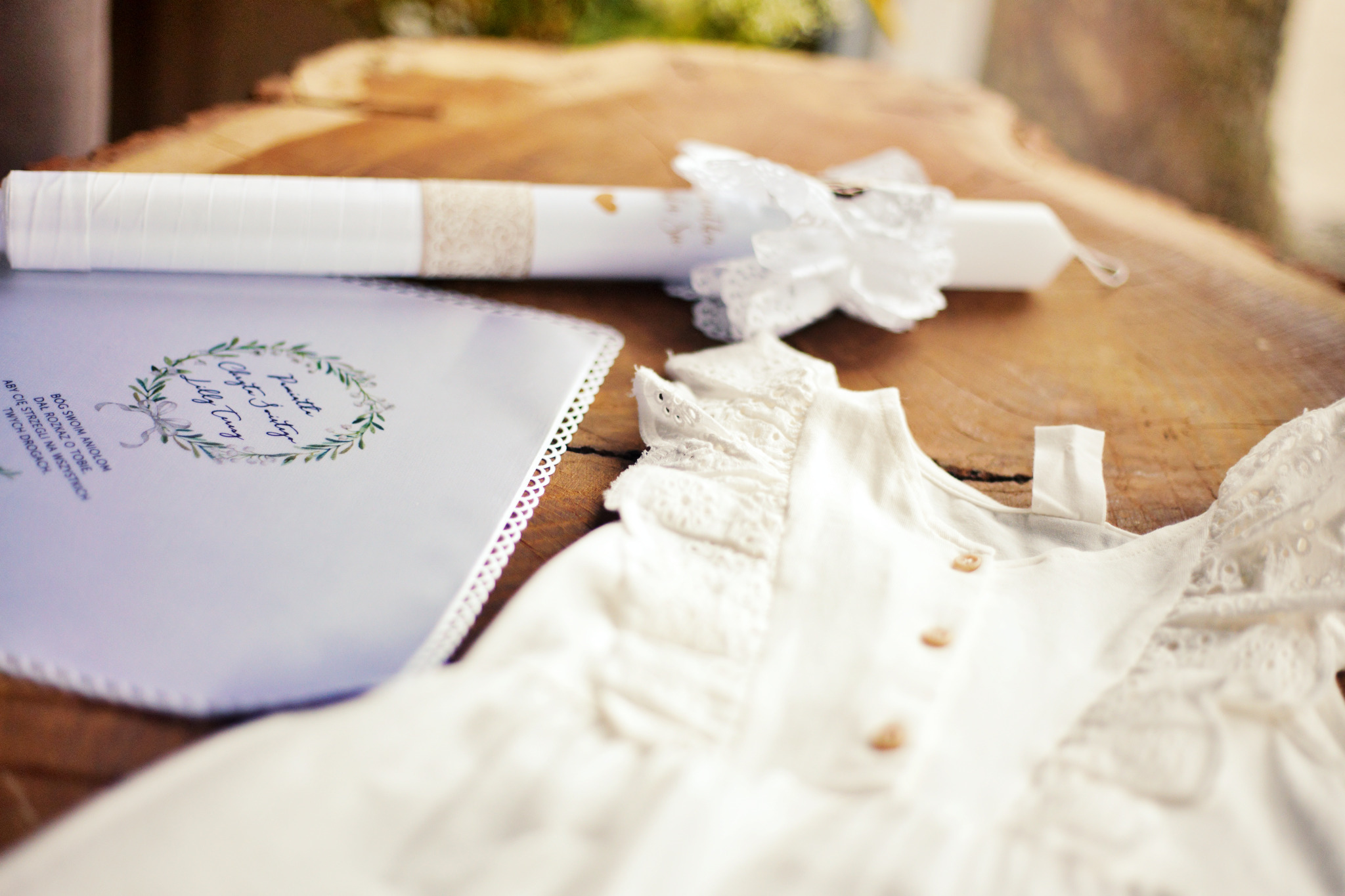 Biała szatka, świeca z koronkową dekoracją i zaproszenie na chrzest leżą na drewnianym blacie, widoczne detale haftu i napisy na zaproszeniu.