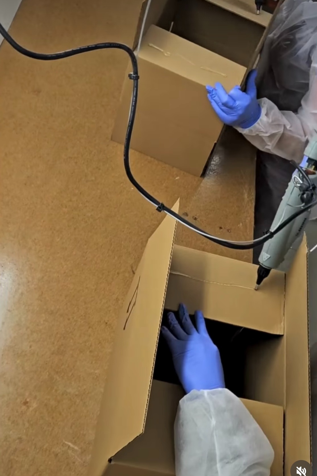 Pracownik w kombinezonie ochronnym i niebieskich rękawicach klei kartonowe pudełko za pomocą pistoletu do kleju na gorąco, prawdopodobnie w Ciechanowie.