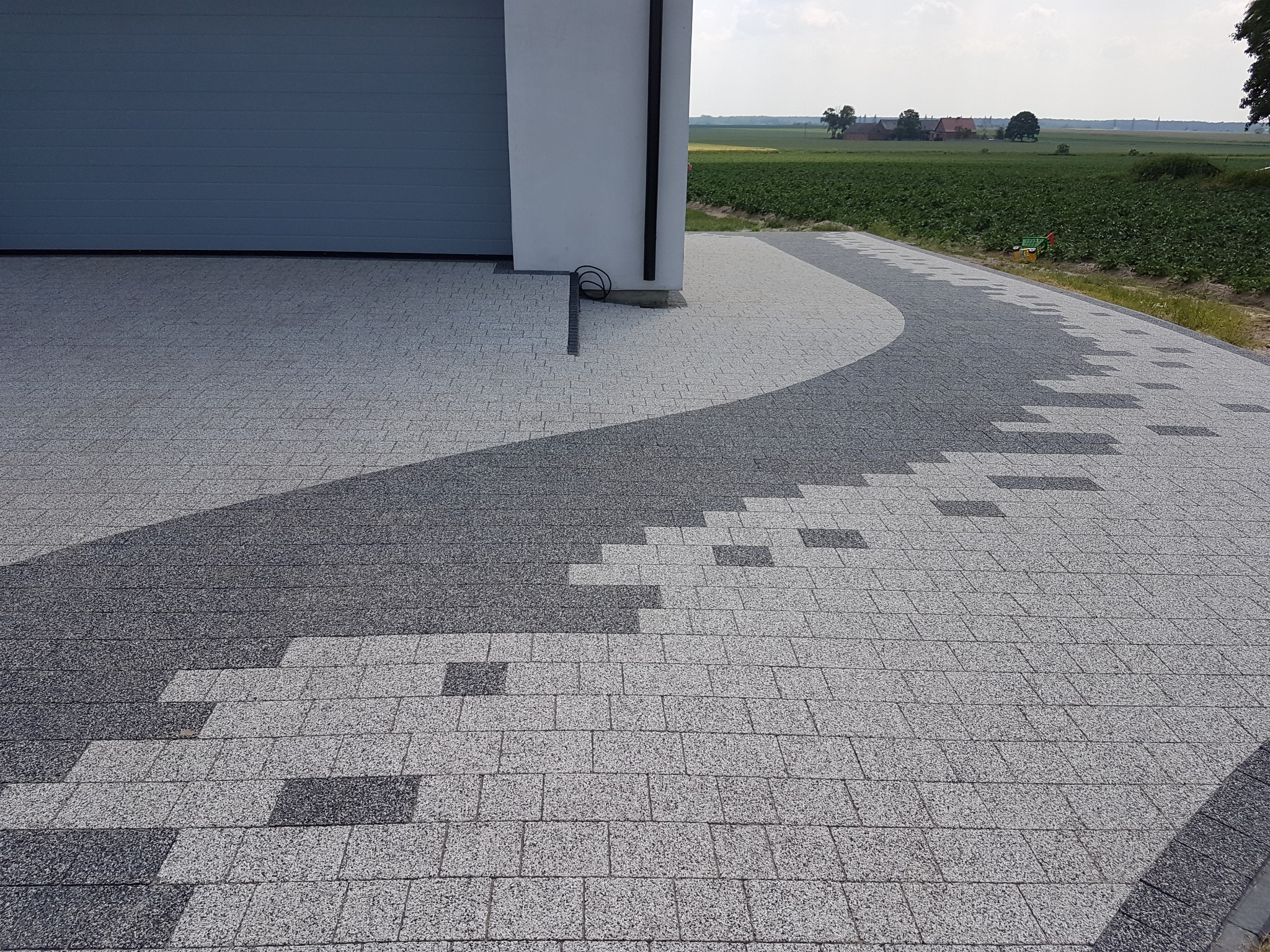 Ułożona kostka brukowa w odcieniach szarości tworząca wzór przed budynkiem z szarą bramą garażową; widoczne pole w tle.