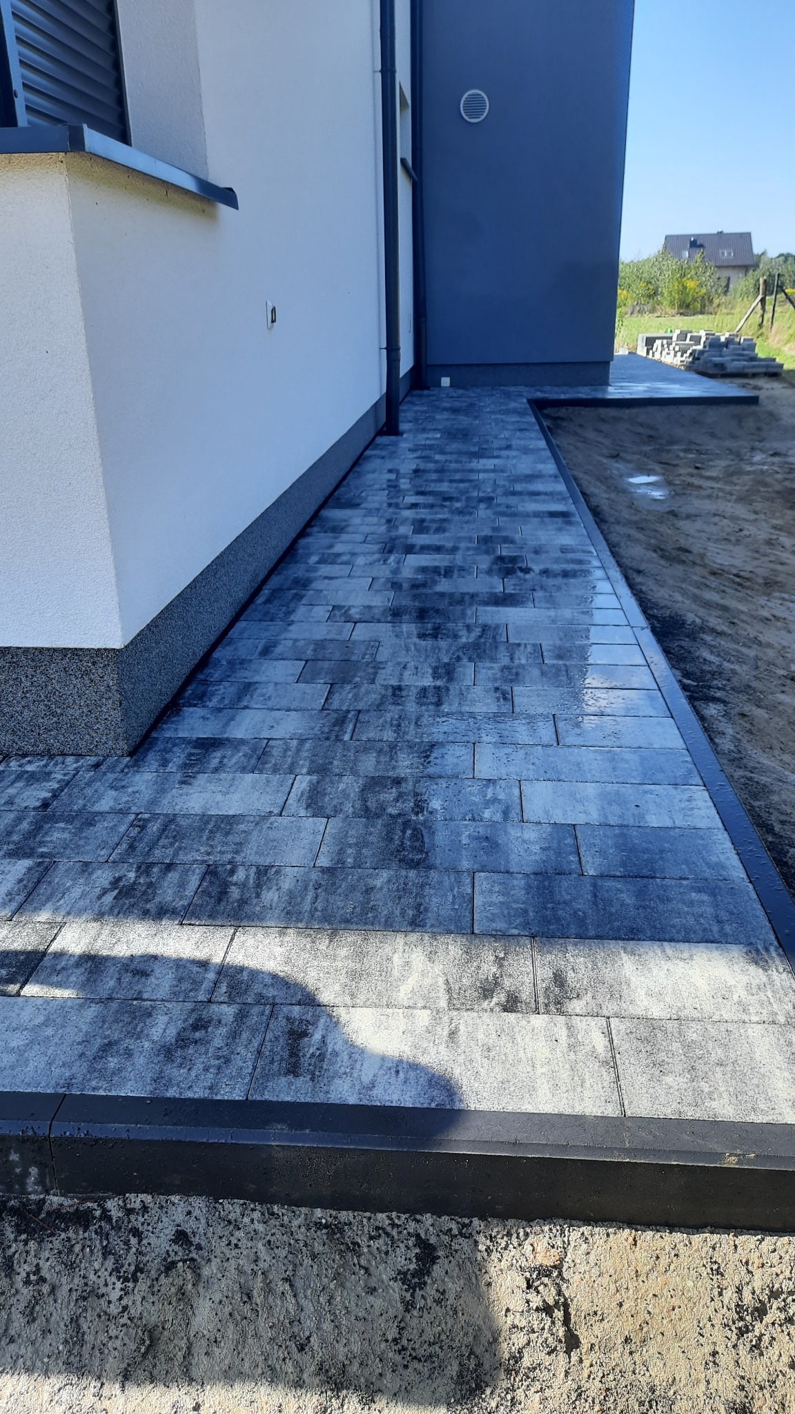 Ułożona ścieżka z szarej kostki brukowej prowadząca wzdłuż ściany budynku, z widocznymi krawężnikami i pozostałościami piasku po pracach.