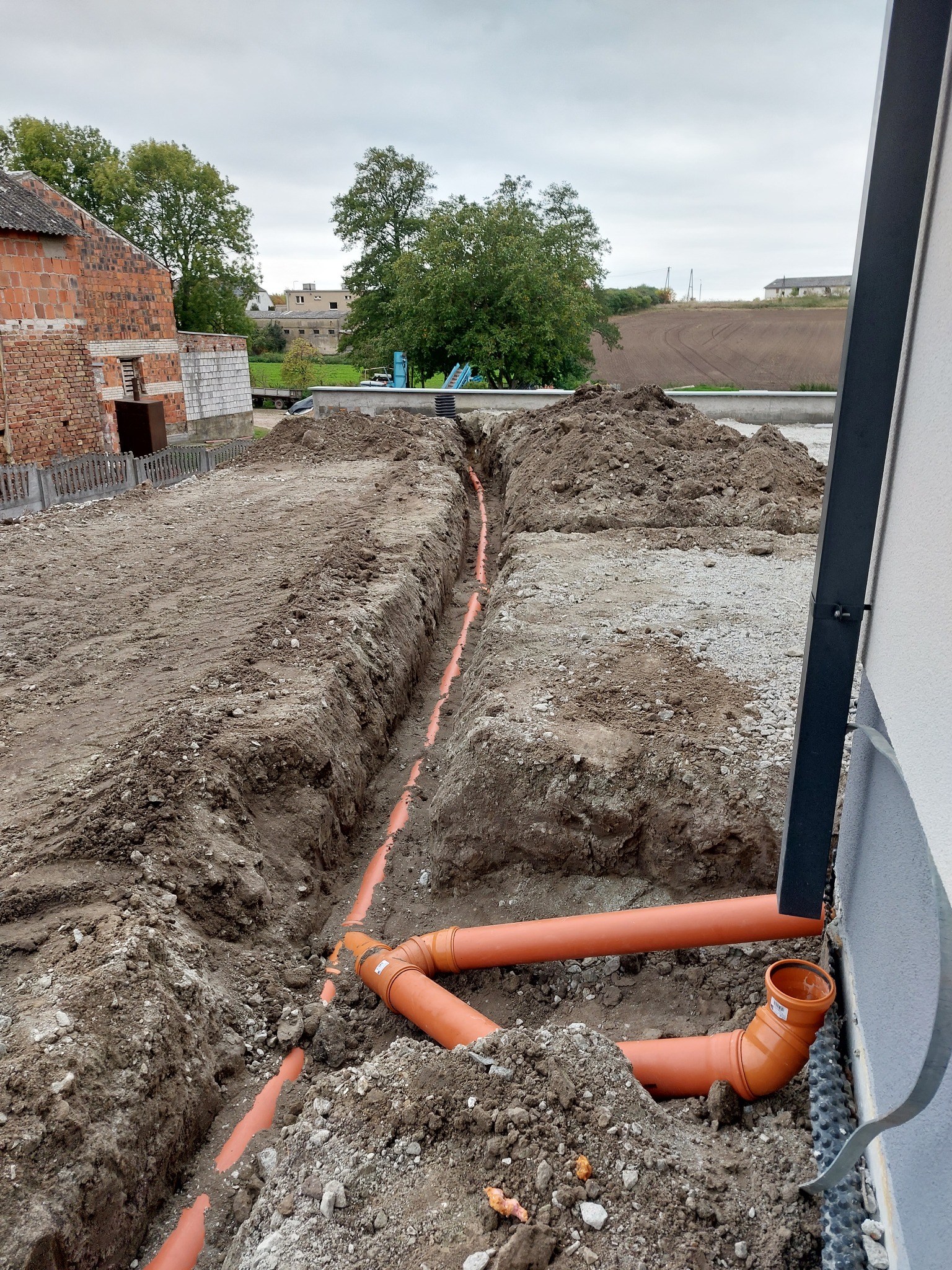 Wykop z pomarańczowymi rurami kanalizacyjnymi ułożonymi w ziemi, widoczne połączenia i nasypy ziemi w tle zabudowa wiejska.