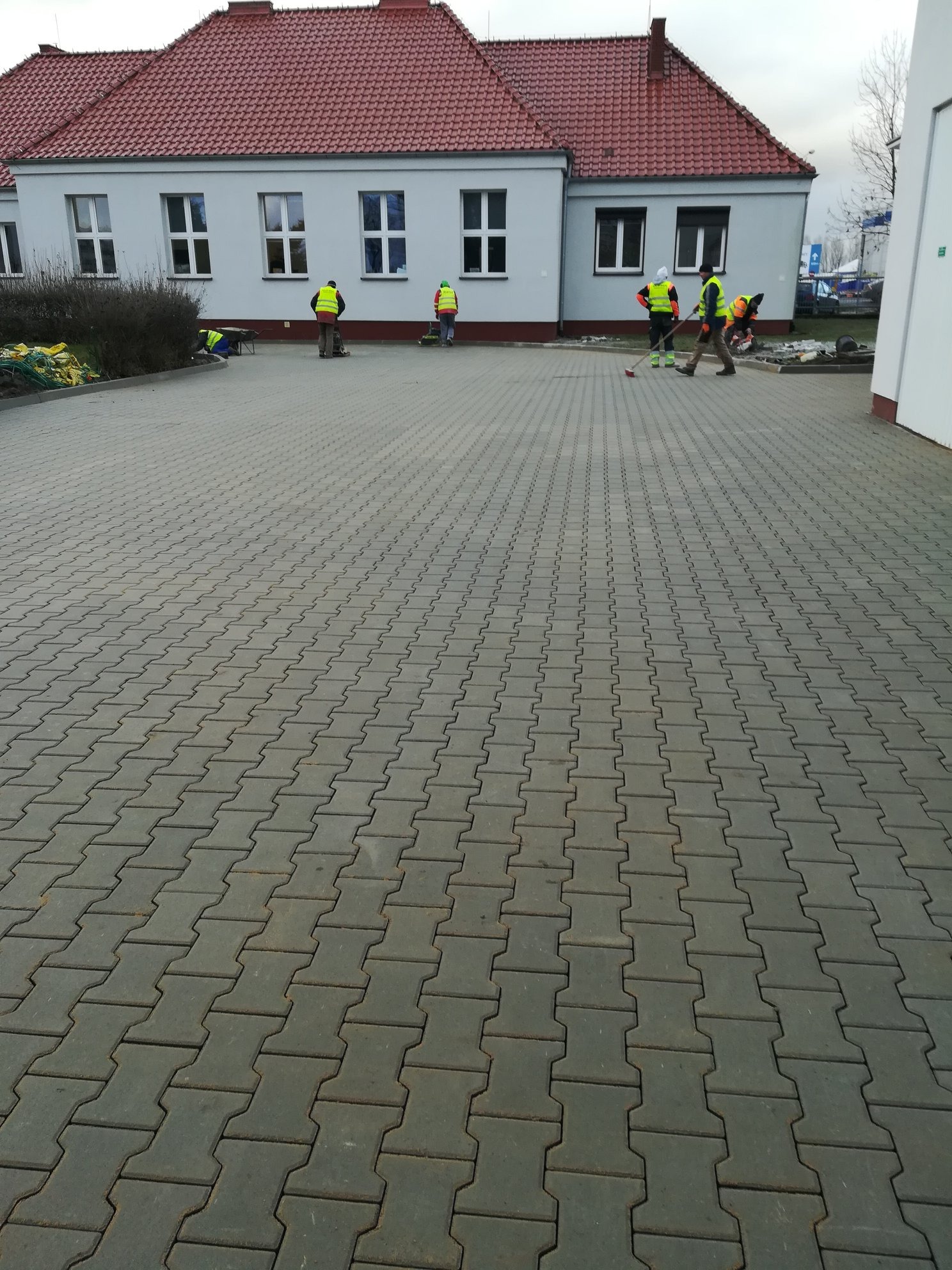 Pracownicy w odblaskowych kamizelkach układają szarą kostkę brukową typu 'I', budynek w tle z czerwonym dachem.