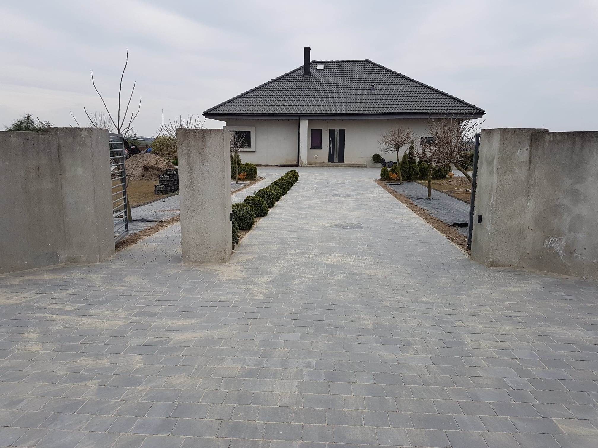 Nowoczesny dom jednorodzinny z szarą kostką brukową na podjeździe, widok przez betonową bramę wjazdową, zieleń ozdobna po bokach podjazdu.