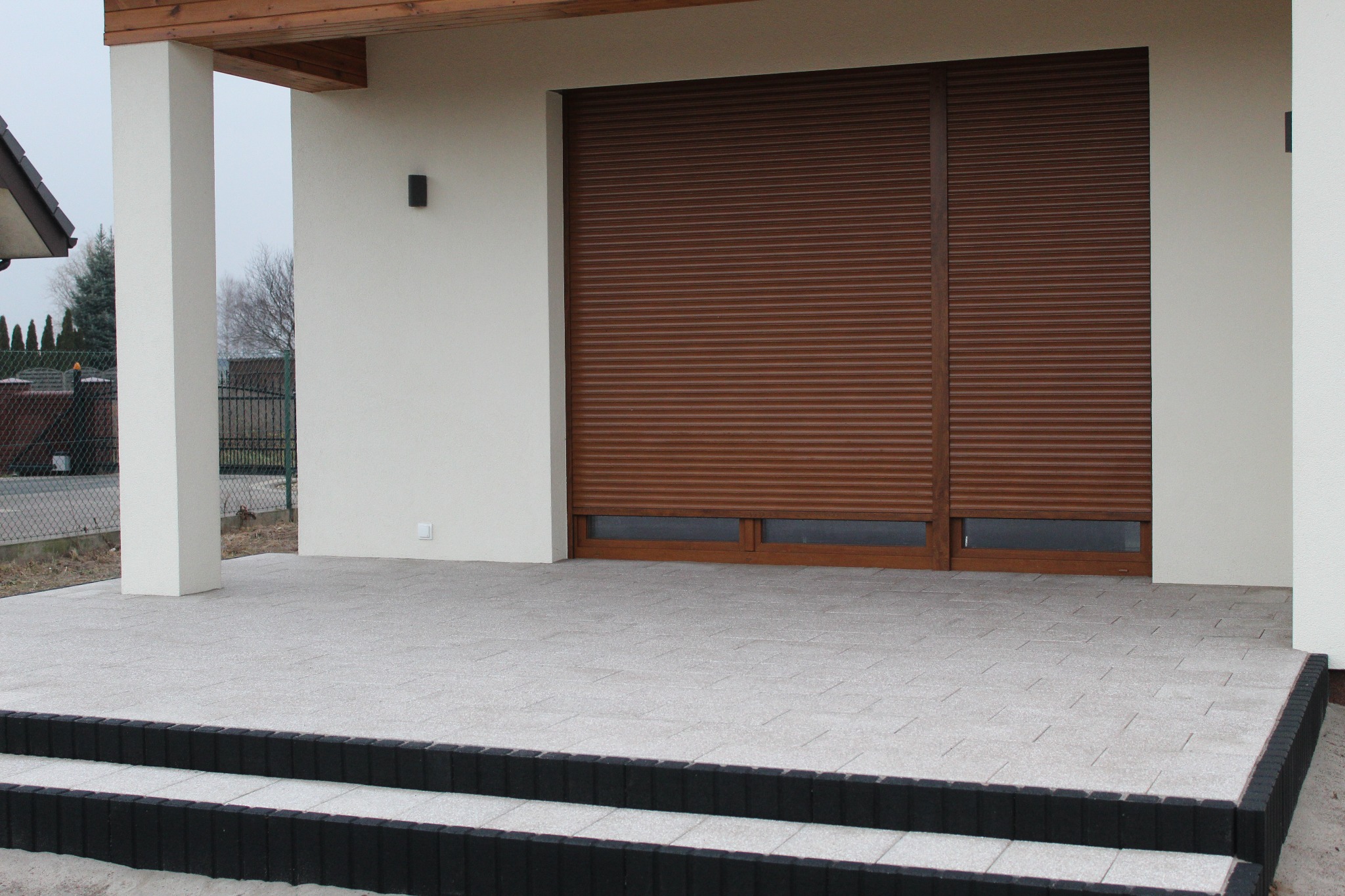 Ułożona kostka brukowa na tarasie przed domem z brązowymi roletami, widoczne czarne obrzeża i dwa stopnie schodów.