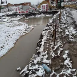 Przygotowanie terenu pod budowę domu: wykopane fundamenty zalane wodą, w tle materiały budowlane i mała koparka.