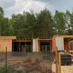 Wznoszenie ścian parteru z czerwonej cegły na budowie domu jednorodzinnego, widoczne elementy szalunków i zbrojenia, w tle las i pochmurne niebo.