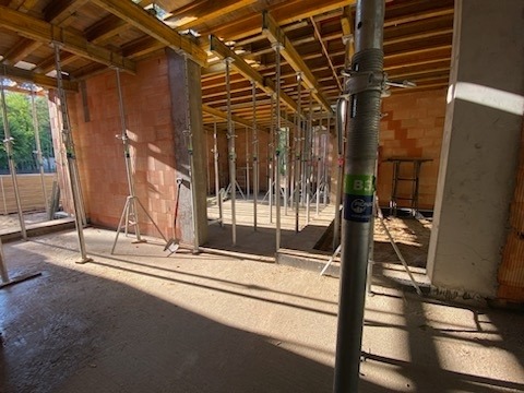 Wnętrze powstającego budynku z czerwonej cegły, widoczne podpory stropowe, częściowo zalana posadzka w cieniu i słońcu.