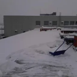 Śnieg na dachu budynku z częściowo odśnieżoną powierzchnią i niebieską łopatą do śniegu opartą o ceglany murek, pochmurne niebo w tle.
