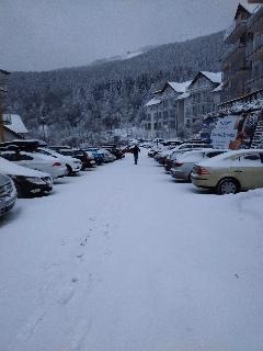 Zasypany śniegiem parking z rzędami samochodów, w tle budynki mieszkalne i zalesione wzgórze, pochmurne niebo i ślady stóp na śniegu.