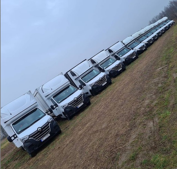 Flota białych busów Renault Master z zabudową kontenerową, ustawionych w rzędzie na nierównym terenie, widok z lekkiego ukosa w pochmurny dzień.