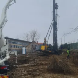 CFA piling rig on a muddy construction site, with a concrete pump truck visible in the foreground and a portable toilet in the background.