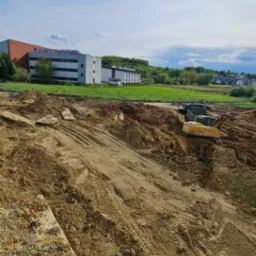 Żółta koparka Atlas 1505 M na placu budowy, widoczne ślady opon na ziemi, w tle budynek Uniwersytetu Rzeszowskiego i zielone wzgórza pod błękitnym niebem.