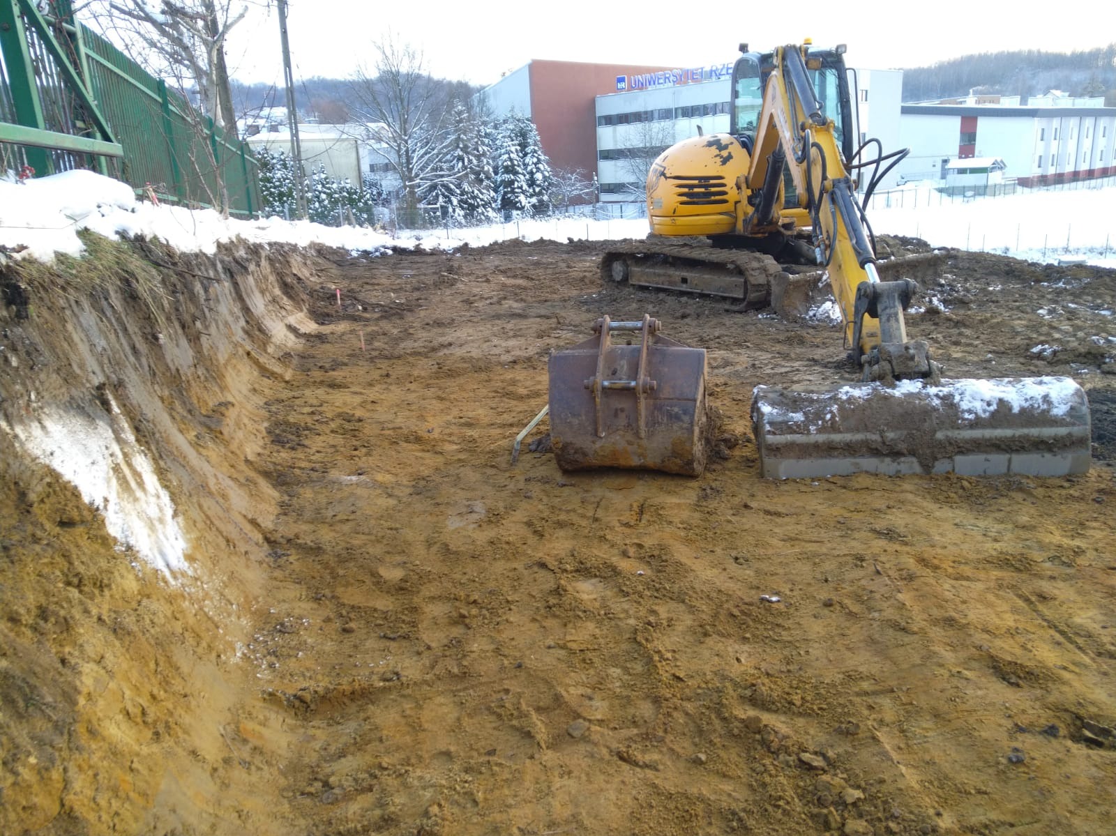 Żółta koparka gąsienicowa z dwoma łyżkami na placu budowy w zimowej scenerii, widoczny wykop w ziemi z resztkami śniegu.