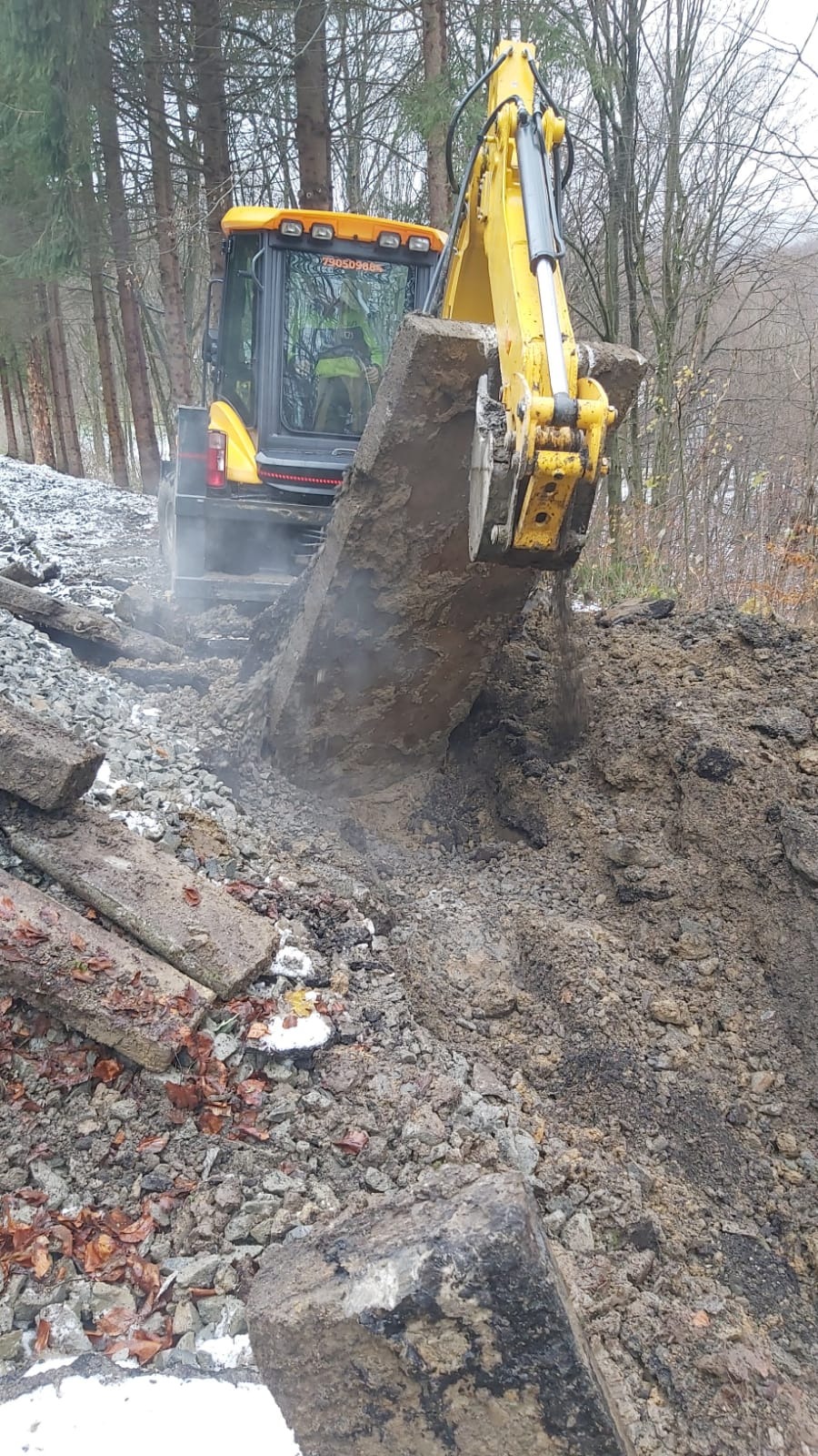 Żółta koparka podnosi betonowy element na nierównym terenie z ziemią i gruzem, w tle widoczne drzewa.