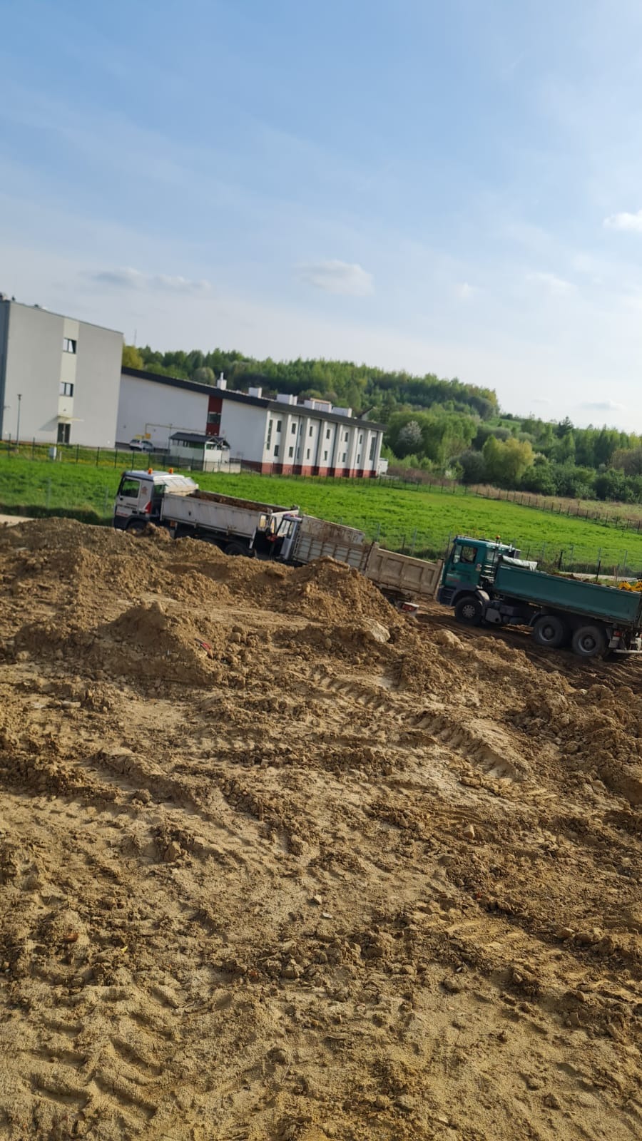 Dwa samochody ciężarowe wyładowują ziemię na placu budowy; w tle budynki i zielone wzgórza pod błękitnym niebem.