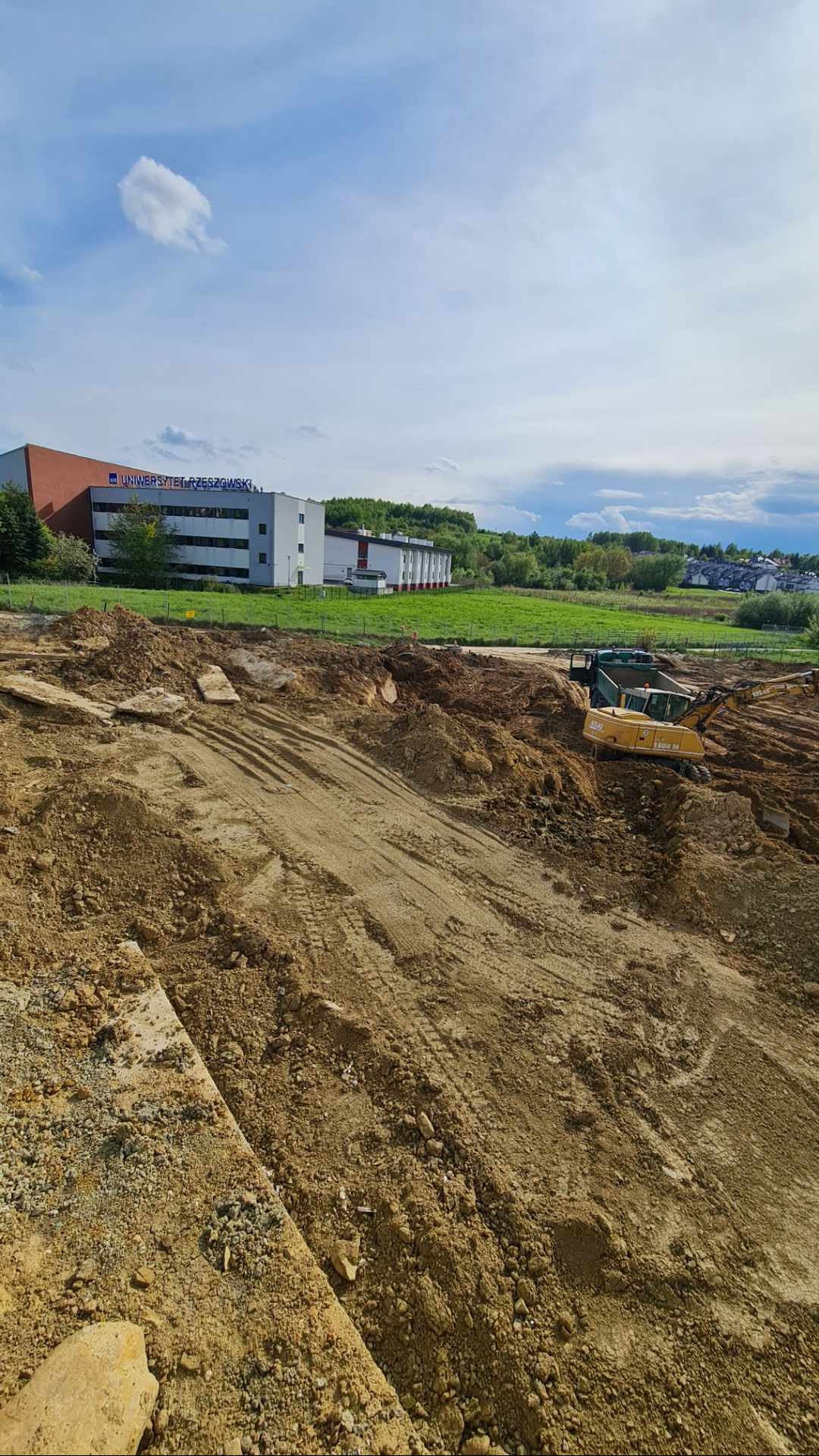 Żółta koparka Atlas 1505 M na placu budowy, widoczne ślady opon na ziemi, w tle budynek Uniwersytetu Rzeszowskiego i zielone wzgórza pod błękitnym niebem.