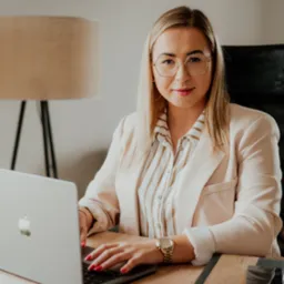 Blondynka w okularach i beżowym żakiecie, siedząca przy biurku z laptopem, na którym widać logo Apple; obok leży wizytówka i notatnik.