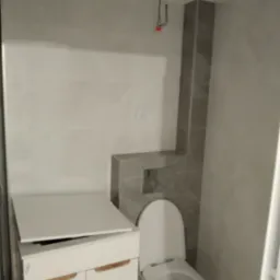 Nowa, częściowo ukończona łazienka z białą szafką pod umywalkę, toaletą z podtynkowym systemem spłukiwania obudowanym szarymi płytkami i wiszącymi kablami oświetleniowymi.