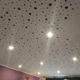 Biały sufit podwieszany z perforacją i wbudowanymi punktowymi lampami LED, widok z dołu, fragment fioletowej ściany w tle.