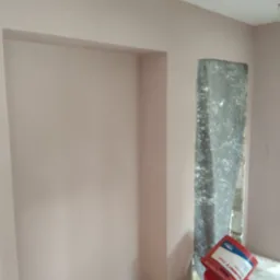 Świeżo położona, jasnoróżowa gładź szpachlowa na ścianie z wnęką i ościeżnicą, widoczne wiadro z masą szpachlową 'Schnitt 6060' i inne akcesoria malarskie.