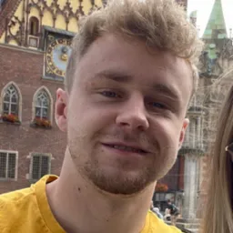 Portret uśmiechniętego mężczyzny z blond włosami na tle zabytkowej architektury Wrocławia. Ujęcie z bliska, widoczna część osoby towarzyszącej po prawej stronie.
