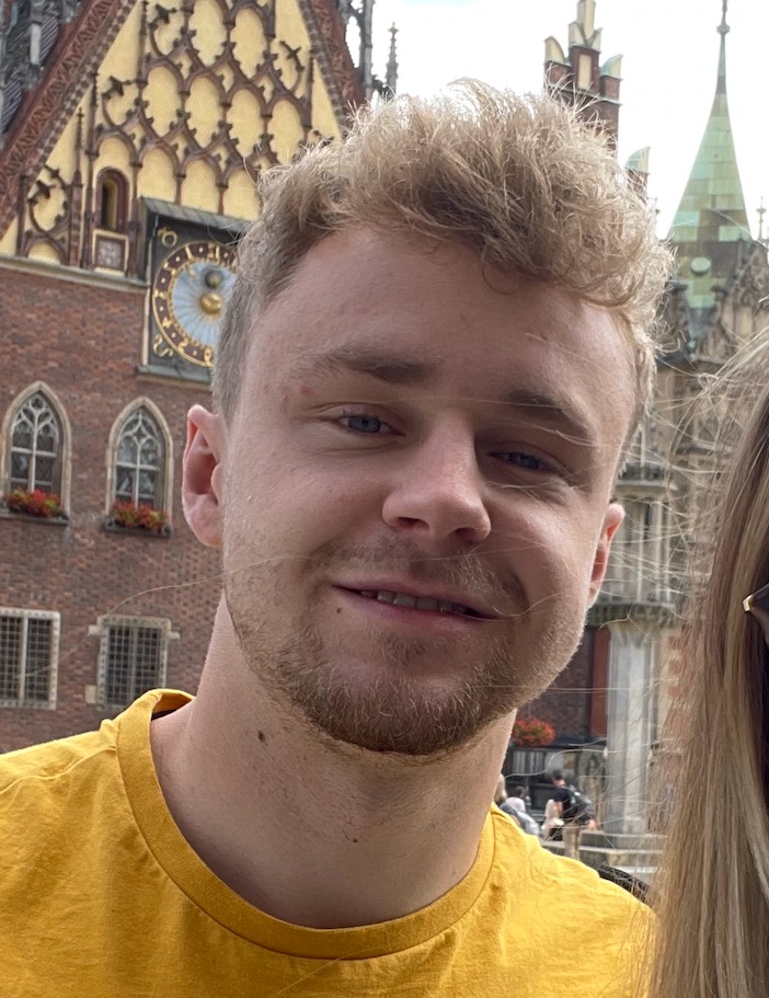 Portret uśmiechniętego mężczyzny z blond włosami na tle zabytkowej architektury Wrocławia. Ujęcie z bliska, widoczna część osoby towarzyszącej po prawej stronie.