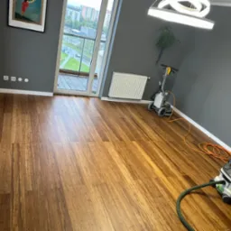 Pokój z parkietem bambusowym w trakcie renowacji. Widoczny odkurzacz przemysłowy i szlifierka do podłóg, balkon z widokiem na zieleń miejską, szare ściany.