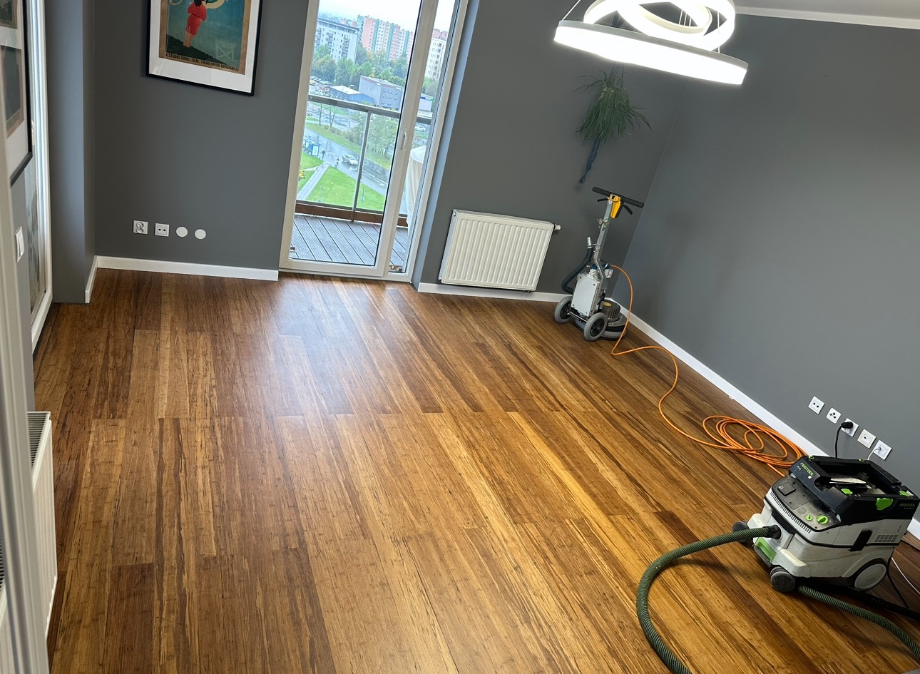 Pokój z parkietem bambusowym w trakcie renowacji. Widoczny odkurzacz przemysłowy i szlifierka do podłóg, balkon z widokiem na zieleń miejską, szare ściany.