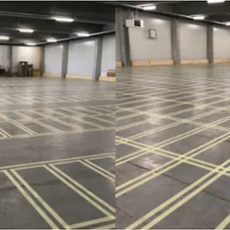 Oznakowanie podłogi w hali magazynowej, żółte linie wyznaczają strefy, w tle widoczne pojemniki i sprzęt.