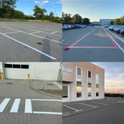 Oznakowanie poziome parkingu z wyznaczonymi miejscami postojowymi, pasami ruchu w kolorze białym i czerwonym, widoczne budynki w tle, oraz pomarańczowe pachołki.