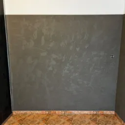 Szara ściana z efektem dekoracyjnym, biały sufit, brązowe płytki podłogowe w romby, ciemne drzwi po lewej stronie, widoczne gniazdko elektryczne.