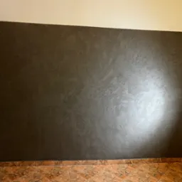 Ściana w kolorze grafitowym z dekoracyjną fakturą, oświetlona lampką biurkową, z widoczną podłogą wyłożoną brązowymi płytkami.