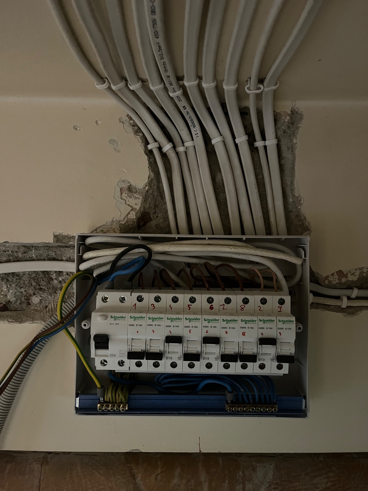 Rozdzielnica elektryczna z przewodami w ścianie, widoczne bezpieczniki automatyczne Schneider Electric, instalacja w trakcie montażu, ściana z ubytkami tynku.