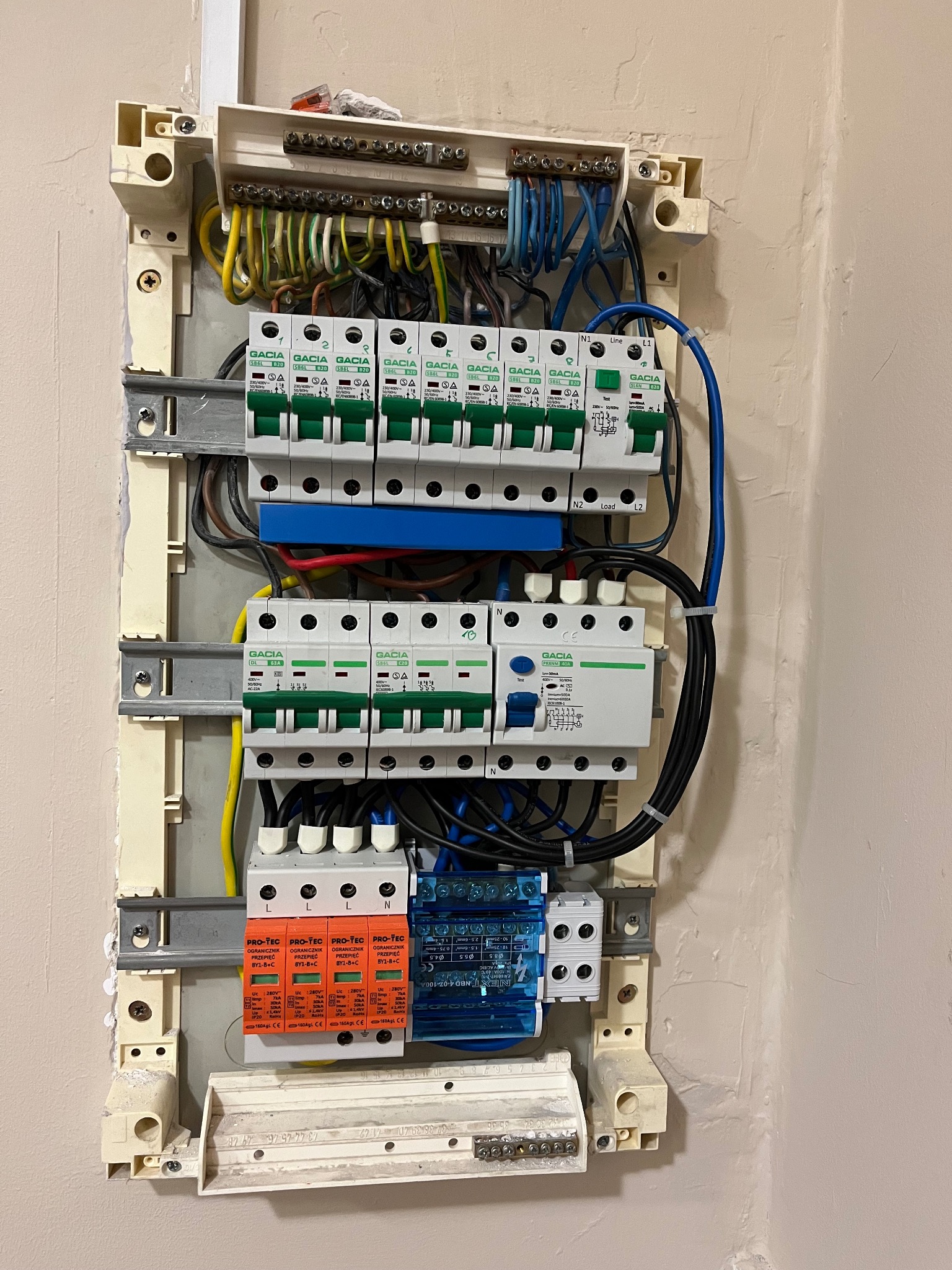 Otwarta skrzynka elektryczna z bezpiecznikami, ogranicznikami przepięć i przewodami różnego koloru, zamontowana na jasnej ścianie, widoczne elementy instalacji elektrycznej.