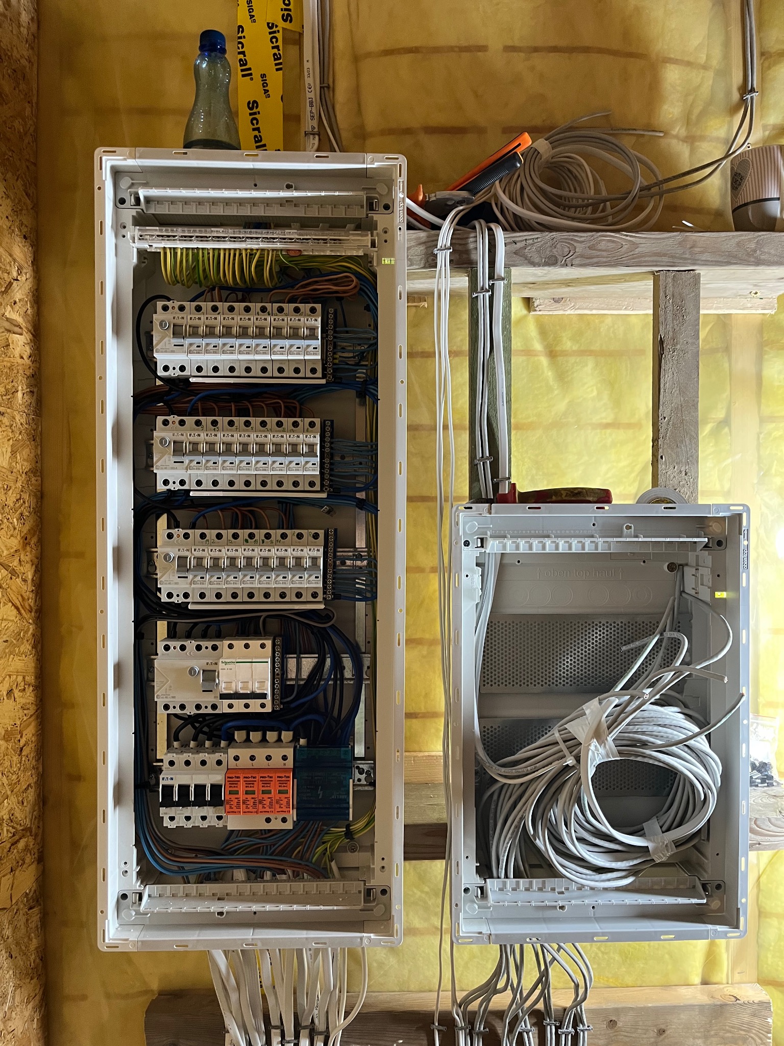 Dwa rozdzielnice elektryczne w trakcie montażu na ścianie z izolacją. Jedna z zamontowanymi bezpiecznikami i okablowaniem, druga pusta z zwiniętym kablem w środku. Na górze półka z narzędziami...