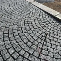 Układanie kostki granitowej w łukowaty wzór, widoczne narzędzie wbite w spoinę, w tle zaparkowany bus i pryzma kostki.