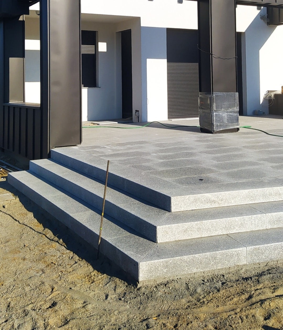 Granitowe schody zewnętrzne prowadzące do tarasu, widoczny pręt pomiarowy wbity w ziemię, w tle nowoczesny budynek z czarnymi elementami konstrukcyjnymi.