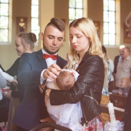 Kinga Górska Fotografia - Rodzice w kościele delikatnie poprawiają czapeczkę niemowlęciu ubranemu w białą sukienkę do chrztu, w tle goście i okna