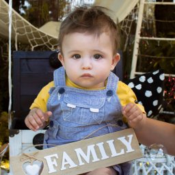 Kinga Górska Fotografia - Uroczy niemowlak w dżinsowych ogrodniczkach i żółtej koszulce siedzi na białym stołku, trzymając drewniany napis 'FAMILY' z wyciętym sercem; w tle dekoracje w stylu rustykalnym i bańki mydlane.