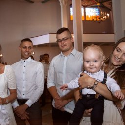 Kinga Górska Fotografia - Uśmiechnięta rodzina pozuje w kościele podczas uroczystości, kobieta trzyma na rękach roześmianego chłopca w eleganckich spodniach na szelkach i koszuli.