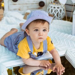 Kinga Górska Fotografia - Uroczy niemowlak w paski kapeluszu i żółtej koszulce leży na brzuchu, opierając dłonie na lustrzanym blacie, z odbiciem w lustrze, w tle białe skrzynie i dekoracje.