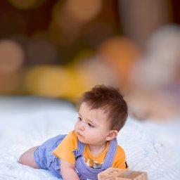 Kinga Górska Fotografia - Siedzący na brzuchu niemowlak w dżinsowych ogrodniczkach i żółtej koszulce, obok drewnianego klocka z datą '08 November', na białym, puszystym kocyku, z rozmytym tłem.