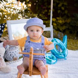 Kinga Górska Fotografia - Uroczy niemowlak w niebieskim kapeluszu i ogrodniczkach siedzi na drewnianym koniku na biegunach podczas sesji zdjęciowej na białym kocu z dekoracjami, w tle zieleń i biała rama.
