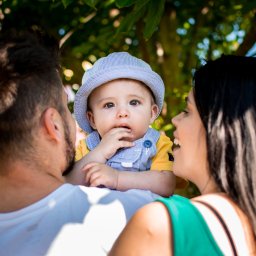 Kinga Górska Fotografia - Uśmiechnięta rodzina: tata trzyma na ramionach małego synka w paskiwanym kapeluszu i żółtej koszulce, mama patrzy na nich z boku, tło rozmyte.