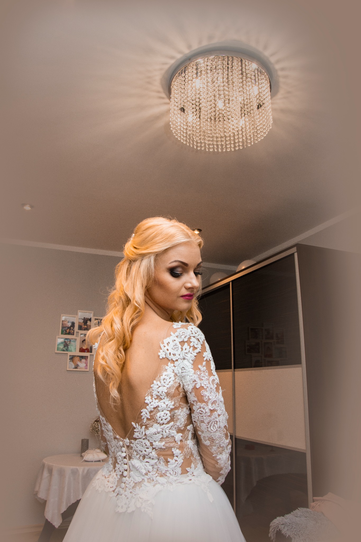 Panna młoda w sukni ślubnej z koronkowymi aplikacjami na plecach, spogląda przez ramię w kierunku światła z kryształowego żyrandola.