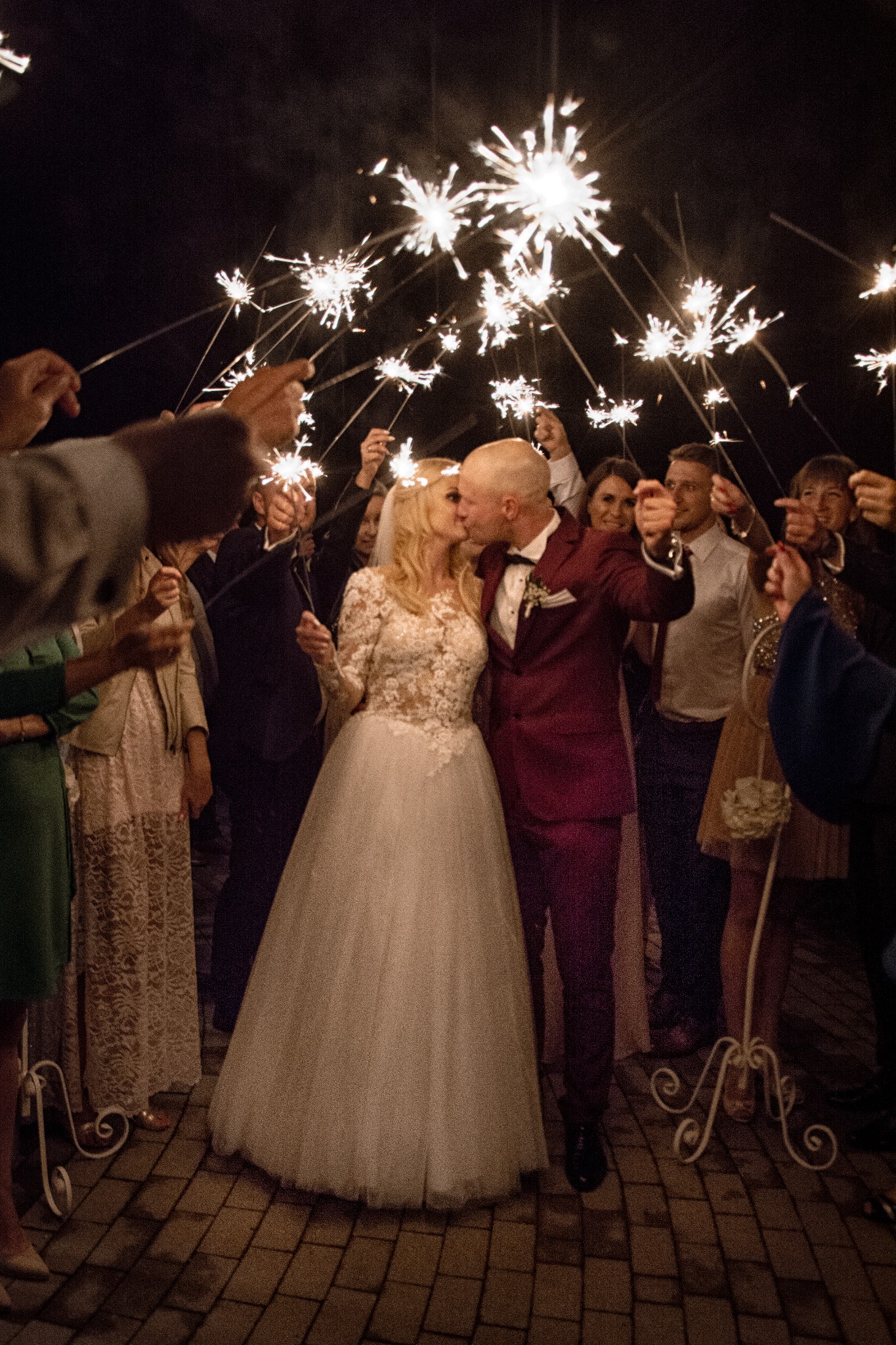 Pocałunek pary młodej w otoczeniu gości trzymających zapalone zimne ognie podczas nocnego przyjęcia weselnego na tle brukowanej nawierzchni.