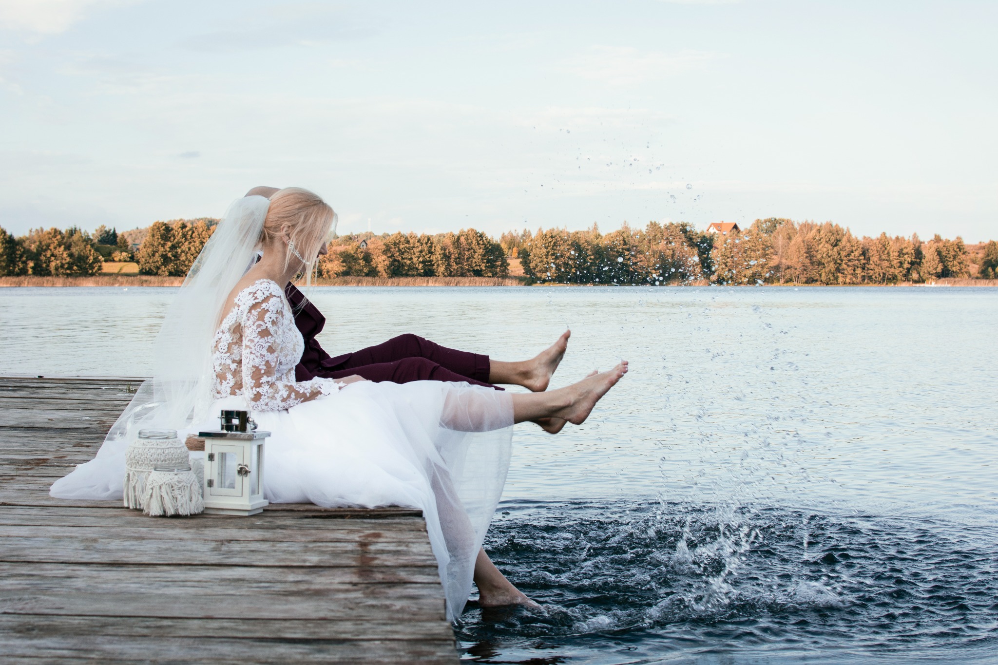 Para młoda siedzi na drewnianym pomoście nad jeziorem, bryzgając wodą stopami; panna młoda w koronkowej sukni i welonie, obok lampiony dekoracyjne.